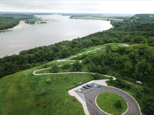 Aerial view of a winding road and parking area near a river, surrounded by lush greenery and rolling hills.