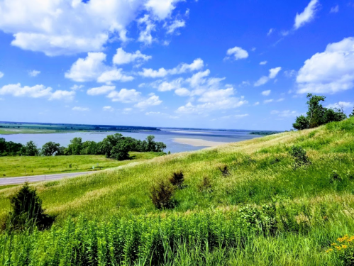 A scenic view of a river with lush green hills and a bright blue sky dotted with clouds.