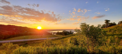 Sunset over a serene landscape with rolling hills, trees, and a calm body of water in the distance.