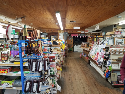 Interior of a rustic general store with shelves stocked with snacks, drinks, and various goods, featuring wooden flooring.