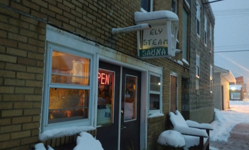 A brick building with a sign reading "Ely Steam Sauna" and an "Open" neon sign, covered in snow.