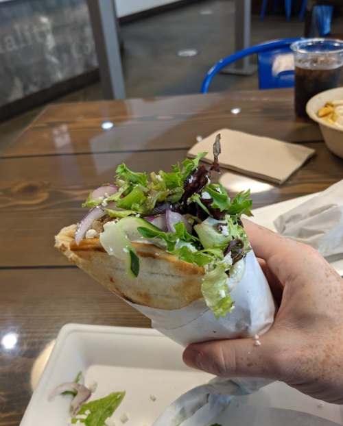 A hand holding a wrapped sandwich filled with greens and meat, with a drink and food container in the background.