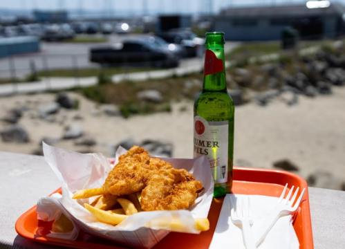 A tray with fish and chips beside a bottle of beer, set against a beachside background.