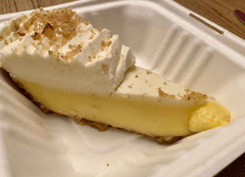 A slice of creamy lemon pie topped with whipped cream and sprinkled with crushed nuts, served in a takeout container.