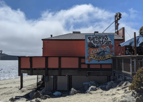 A seaside seafood parlor with a colorful sign, set against a cloudy sky and sandy beach.