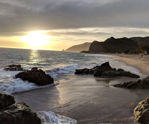 Sunset over a tranquil beach with gentle waves, rocky shoreline, and distant mountains under a cloudy sky.