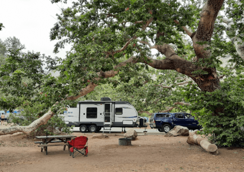 A campsite with a travel trailer, a pickup truck, a picnic table, and a red chair under large trees.