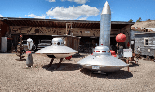 A quirky display of UFO models and alien figures outside a rustic building, with a blue sky and clouds above.