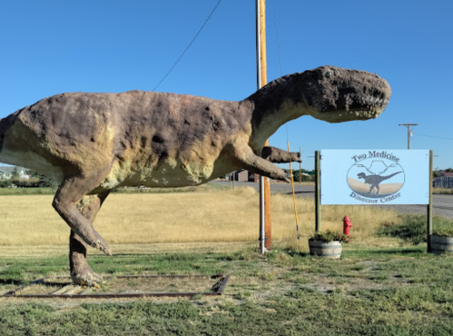 A large dinosaur statue stands outside the Two Medicine Dinosaur Center, with a grassy field in the background.
