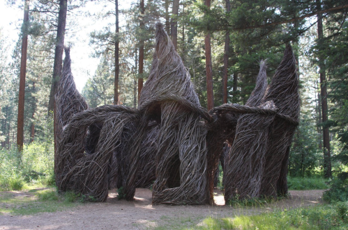 A whimsical structure made of intertwined branches, resembling a forest dwelling, surrounded by tall trees.