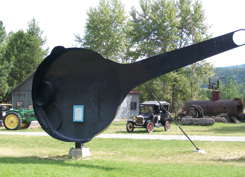A large metal ladle sculpture stands in a grassy area, with vintage vehicles and trees in the background.