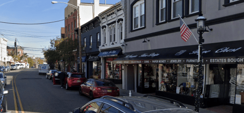 A street view featuring shops with awnings, parked cars, and an American flag on a lamp post.