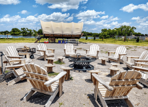 A circle of wooden chairs around a fire pit, with a covered wagon and blue sky in the background.