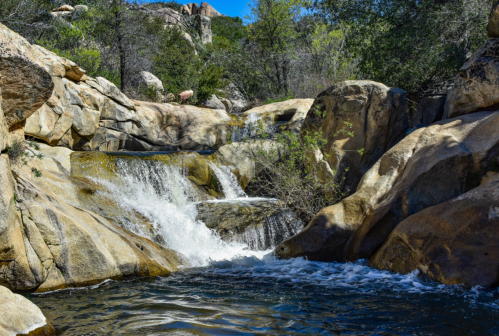 A serene waterfall cascades over rocky terrain into a clear pool, surrounded by lush greenery and boulders.