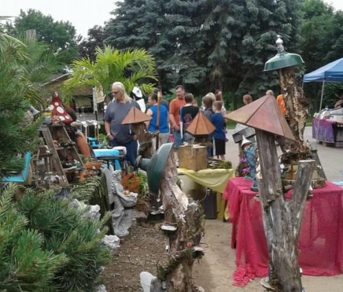 A vibrant outdoor market scene with people browsing unique crafts and decorations among greenery and colorful tables.