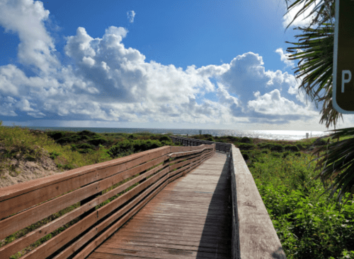 A wooden boardwalk leads to a beach, surrounded by lush greenery and under a bright blue sky with fluffy clouds.
