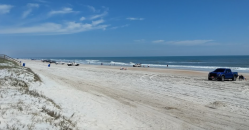 A sandy beach with vehicles parked along the shore, gentle waves, and a clear blue sky.