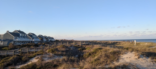 Coastal scene with beach houses, sandy dunes, and a clear blue sky overlooking the ocean.