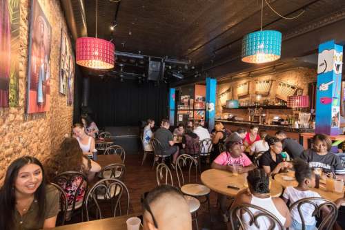 A lively café interior with patrons at tables, colorful lamps, and a bar area, featuring exposed brick walls and artwork.