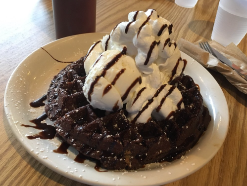 A chocolate waffle topped with whipped cream and drizzled with chocolate sauce, served on a white plate.