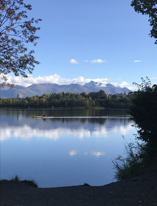 A serene lake reflects mountains and a blue sky, with a lone rower in the distance and trees framing the scene.