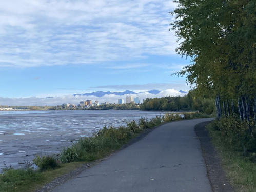 A scenic pathway along a river with city buildings and mountains in the background under a partly cloudy sky.