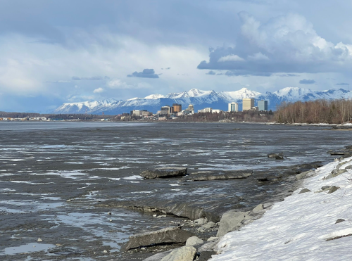 A scenic view of a frozen lake with a city skyline and snow-capped mountains in the background under a cloudy sky.