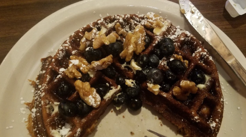 A partially eaten waffle topped with blueberries, walnuts, and powdered sugar on a plate with a knife beside it.