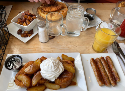 A table with a plate of French toast topped with apples and whipped cream, sausage links, and a side of potatoes.
