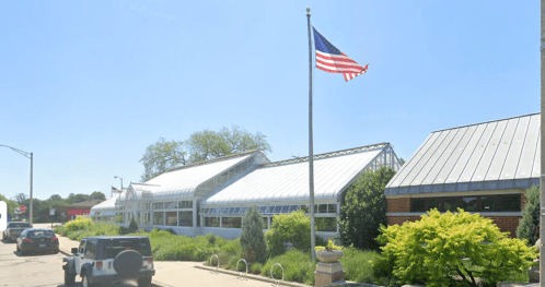 A building with a metal roof and large windows, featuring an American flag in front and greenery surrounding it.