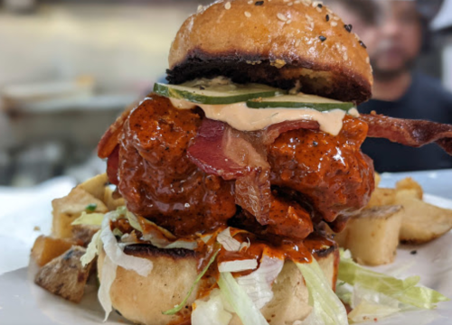 A close-up of a spicy chicken sandwich topped with bacon, lettuce, and sauce, served with crispy potato wedges.
