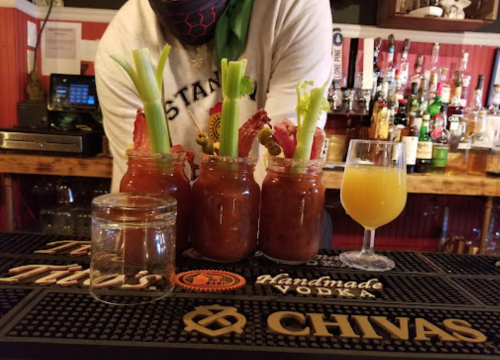Three Bloody Mary cocktails with celery and olives, alongside a glass of orange juice, on a bar counter.