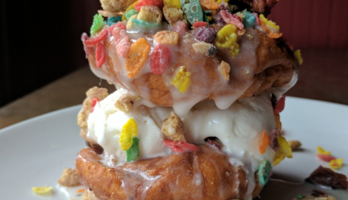 A towering dessert featuring two glazed donuts filled with ice cream and topped with colorful cereal.