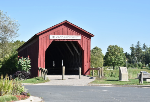 A red covered bridge with a sign, surrounded by greenery and a pathway leading to it.