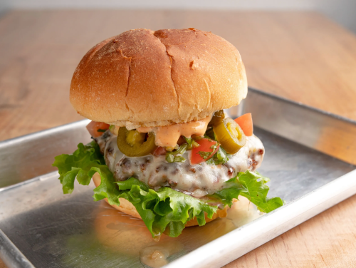 A juicy burger with melted cheese, lettuce, tomatoes, and olives on a toasted bun, served on a metal tray.