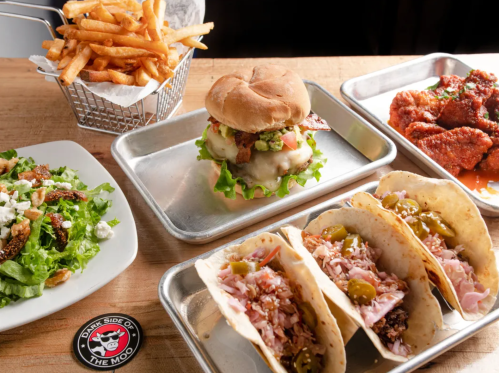 A spread of food including a burger, fries, salad, chicken wings, and tacos on metal trays.