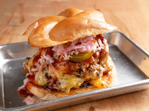 A hearty burger with pulled pork, cheese, pickles, and coleslaw on a toasted bun, served on a metal tray.