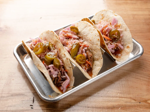 Three tacos on a metal tray, filled with shredded meat, pickled jalapeños, and colorful toppings.