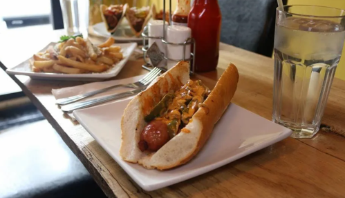 A hot dog in a bun topped with sauce, served on a plate with fries and a glass of water in the background.