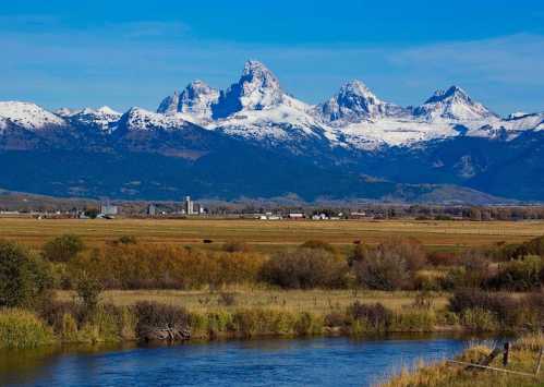 Snow-capped mountains rise above a green valley, with a river and fields in the foreground under a clear blue sky.