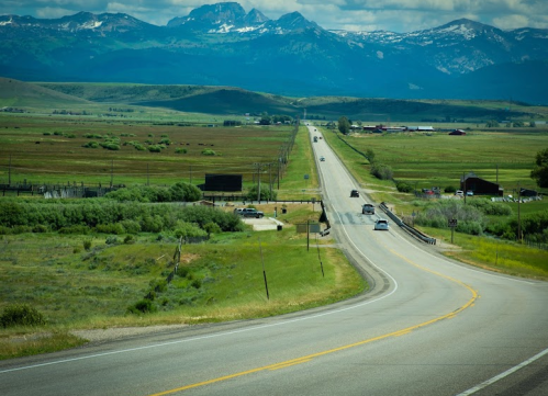 A winding road stretches through green fields, with mountains in the background under a partly cloudy sky.
