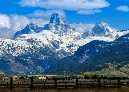 Snow-capped mountains rise majestically under a blue sky, with green valleys and a wooden fence in the foreground.