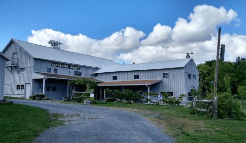 A rustic winery building with a gray exterior, surrounded by greenery and a gravel path under a blue sky with clouds.