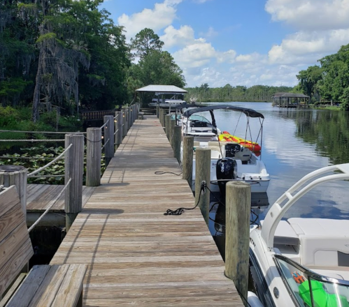 A wooden dock lined with boats, surrounded by lush greenery and calm water under a blue sky with fluffy clouds.