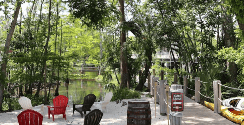A serene outdoor scene with red and black chairs, a sandy area, and lush greenery by a calm waterway.