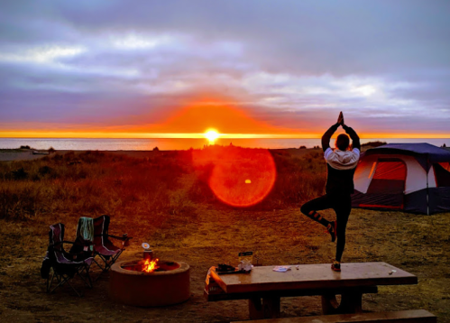 A person practicing yoga at sunset near a campfire, with a tent and chairs in the background.