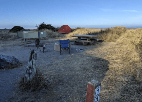 A campsite with two chairs, a picnic table, a fire pit, and a tent in a grassy area under a clear sky.