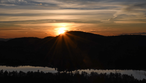 A sunset over a mountain, with rays of light reflecting on a calm lake below.