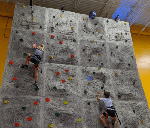 Two children climbing a colorful indoor rock wall with various holds against a bright yellow background.