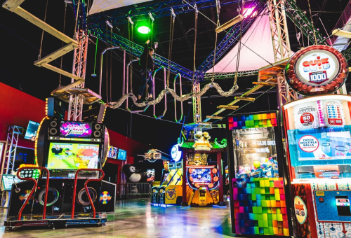 A vibrant arcade with games, colorful lights, and a ropes course overhead, creating a fun atmosphere for visitors.